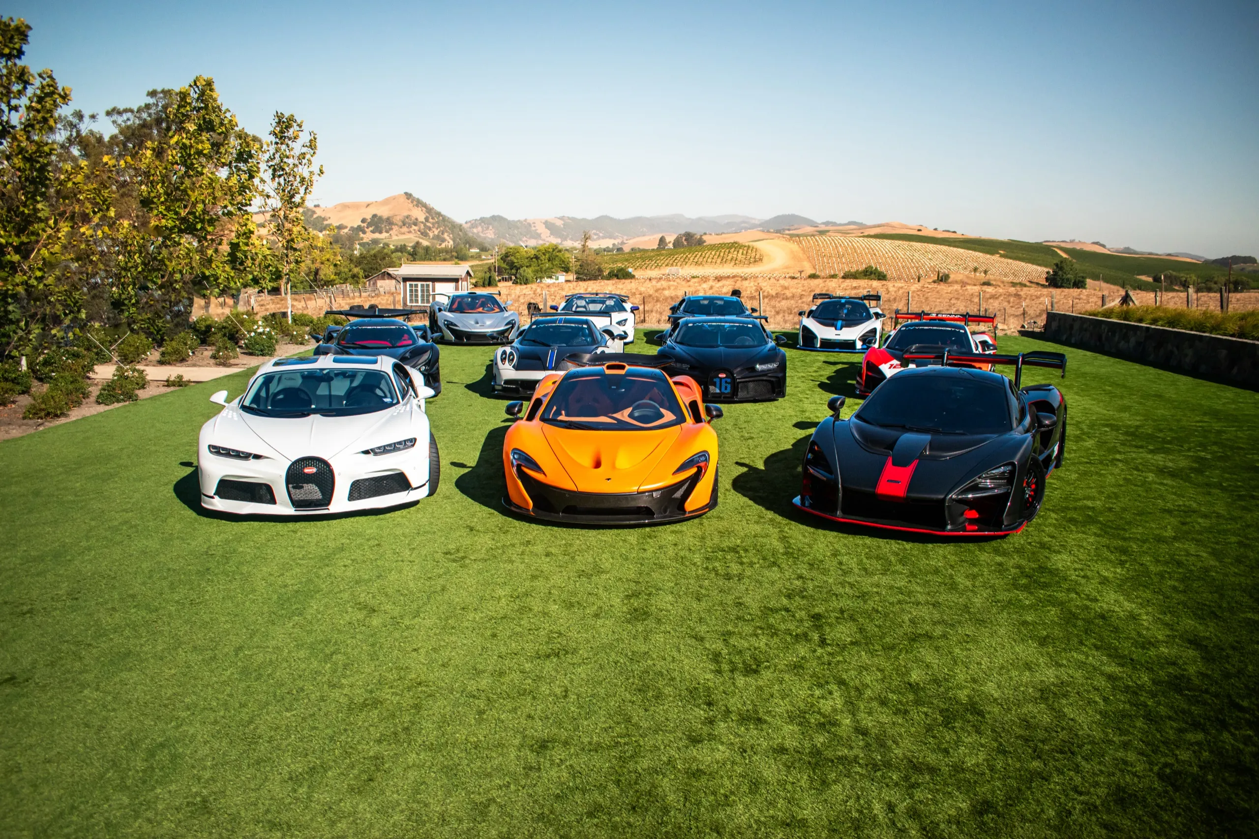 Nine hypercars gathered at a vineyard estate, including a white Bugatti Chiron, an orange McLaren P1 and a McLaren Senna GTR